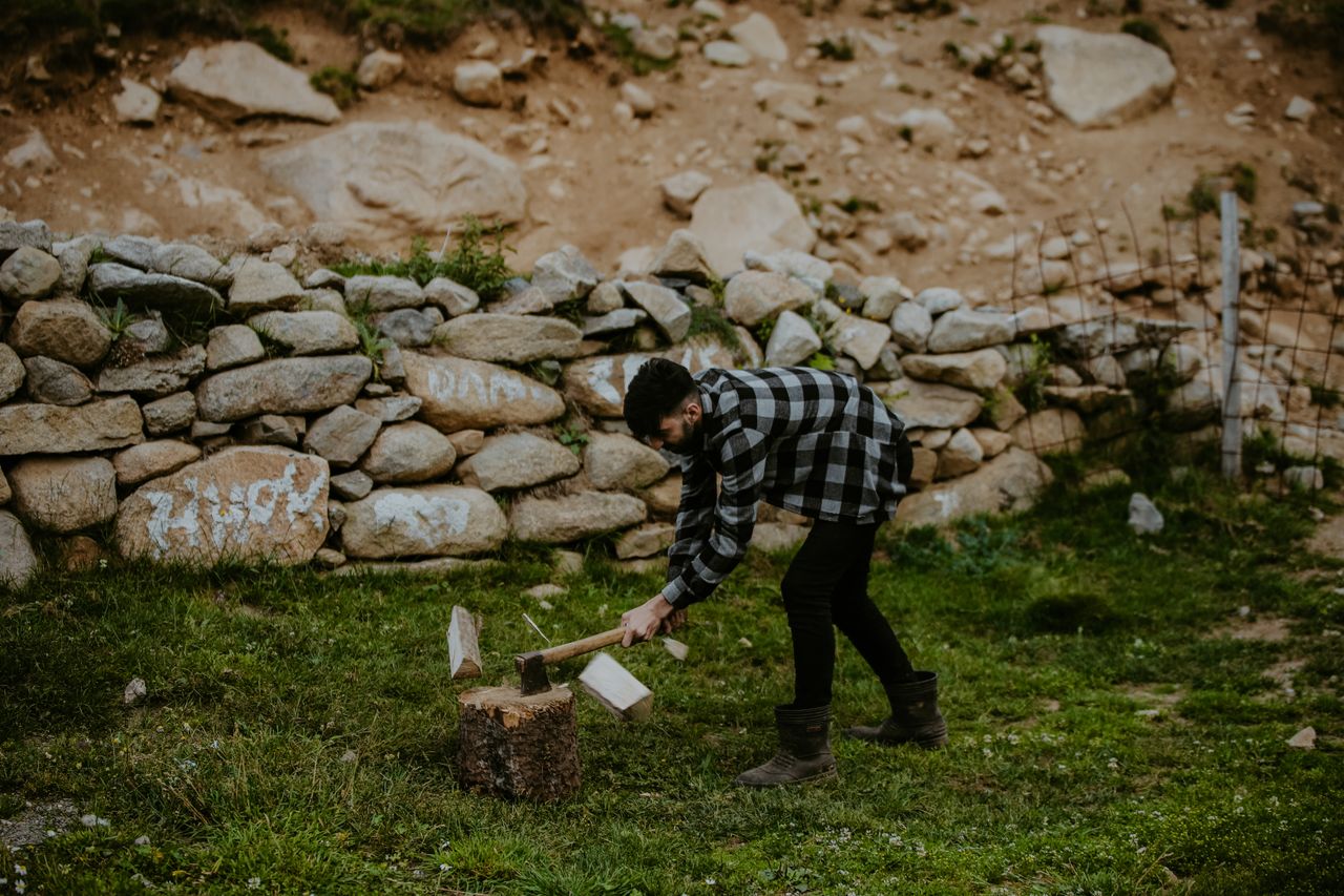 A man chopping wood with an axe next to a stone wall in a rural outdoor setting.